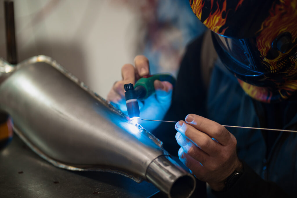 A mechanic in a modern workshop is crouched beside a motorcycle, working on its engine components; tools and parts lie around on the floor and a workbench is visible in the background. Bright overhead lights illuminate the scene, emphasizing metal surfaces and intricate machinery. The image underscores how mechanical repair and maintenance involve sealing systems—like fuel lines, exhausts, or cooling circuits—that must be leak-free, illustrating why leak testing is essential in such manufacturing or post-production settings.