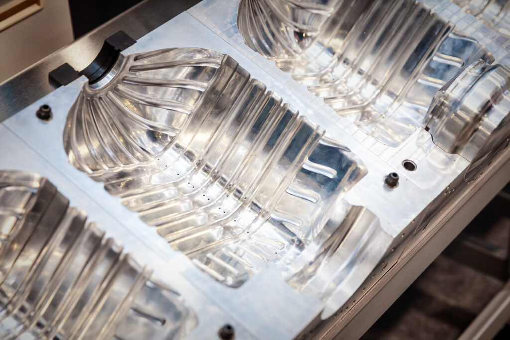 Three translucent molded plastic containers are shown in a tray fixture under bright lighting, suggesting mass production in an injection molding setup; the image highlights molding as a manufacturing process prone to defects such as porosity or leakage. The containers appear uniform in shape, with fine seams and surface details visible. The mold or fixture holding them conveys the industrial context. This visual supports discussion of why molded parts require rigorous leak testing in common manufacturing processes to ensure airtight or watertight integrity.