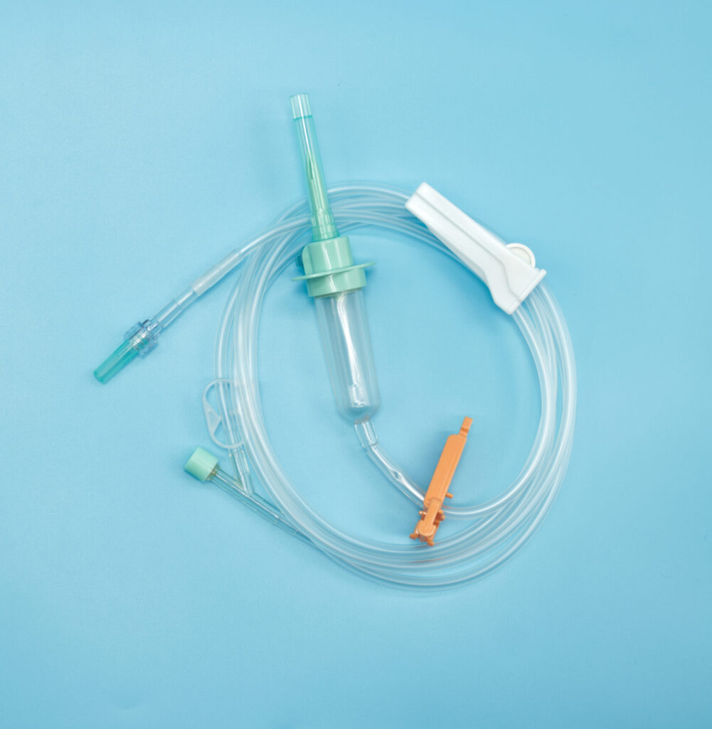A coiled IV set laid on a blue background. It includes a transparent drip chamber, flexible tubing, a green connector, and an orange clamp. These medical-grade plastics are examples of components requiring leak testing.