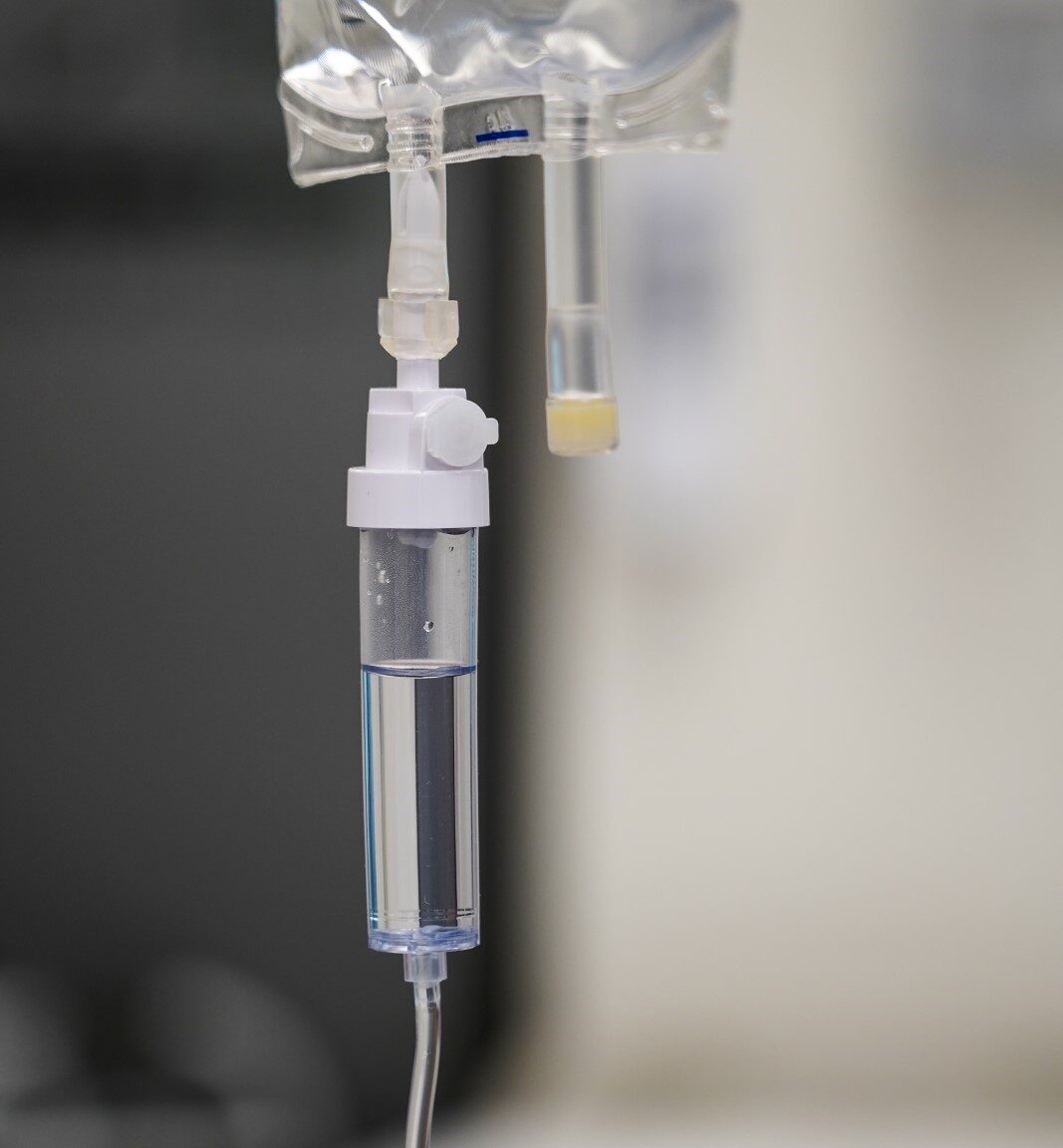 A close-up of a medical IV drip chamber attached to a clear fluid bag, with tubing extending downward for infusion. The transparent chamber shows fluid partially filled, with small air bubbles visible inside. A plastic connector and clamp regulate the flow, while a second line with a capped port hangs beside it. The background is softly blurred, emphasizing the sterile clinical equipment. This image illustrates a precision medical infusion component, the kind of device that requires rigorous leak testing to ensure safe and accurate fluid delivery in healthcare settings.