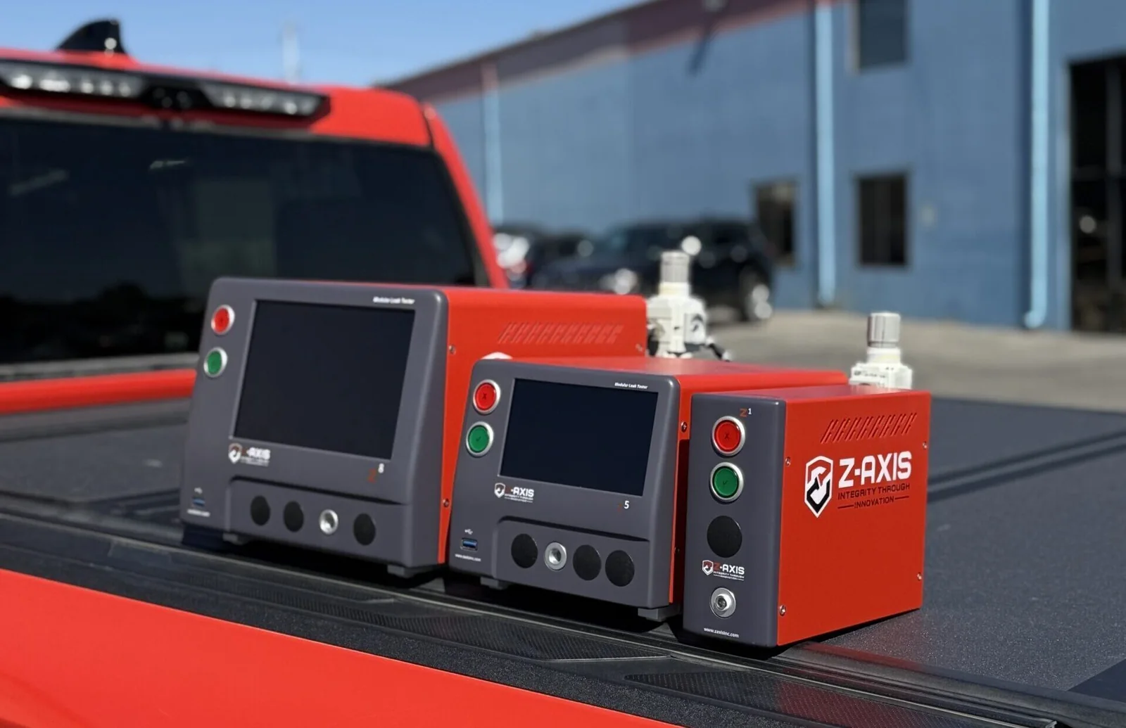 Three Zaxis modular leak testers — the Z1, Z5, and Z8 — are displayed side by side on the back of a red vehicle. Each unit features a large touchscreen, red and green control buttons, and multiple front ports for connections, with the Z-AXIS logo and “Integrity Through Innovation” tagline visible on the red side panels. White fittings are attached on top for air lines. The outdoor industrial setting, with a blue building and parked cars in the background, highlights the rugged, portable design of Zaxis precision air leak detection systems for industrial quality control applications.